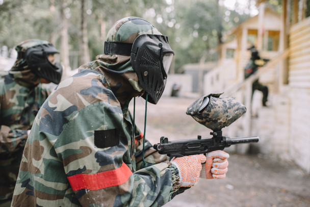 side view of male paintballer and his team in uniform and protective masks aiming by paintball guns outdoors - Foto, imagen