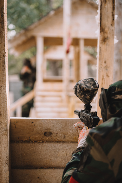 partial view of paintball player in camouflage uniform aiming by paintball gun from wooden tower outdoors  - Photo, Image