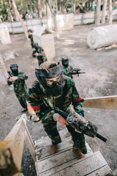high angle view of male paintball player in goggle mask with marker gun on staircase while his team standing behind outdoors  - Fotografie, Obrázek