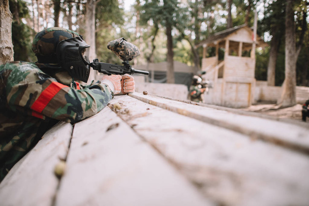 selective focus of paintball player in protective mask and camouflage aiming by marker gun outdoors  - Foto, afbeelding
