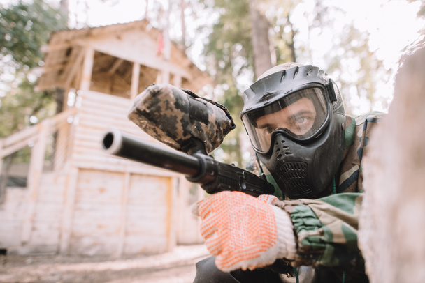 serious male paintball player in goggle mask and camouflage aiming by paintball gun outdoors  - 写真・画像