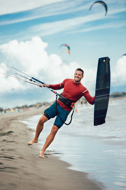 Bonito homem caucasiano surfista profissional em pé na praia de areia com seu papagaio e placa
. - Foto, Imagem
