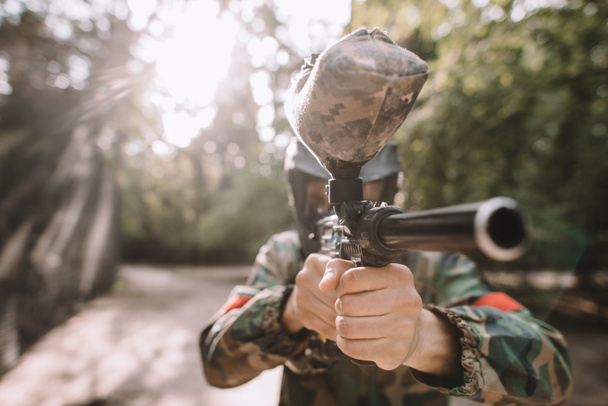selective focus of male paintball player in goggle mask and camouflage aiming by paintball gun outdoors  - Photo, Image