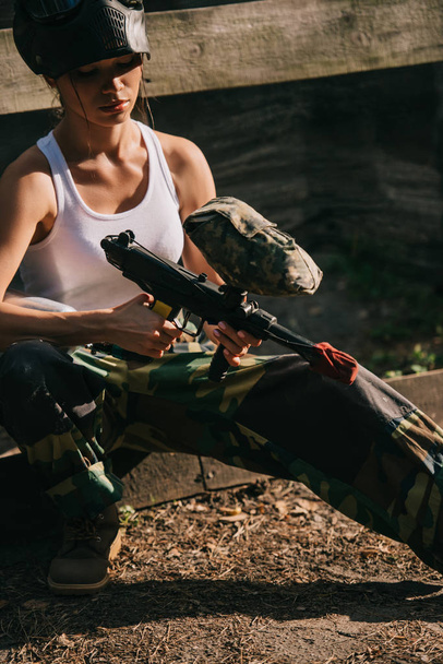 geconcentreerd vrouwelijke paintballer in witte singlet en goggle masker laden paintball pistool buitenshuis - Foto, afbeelding