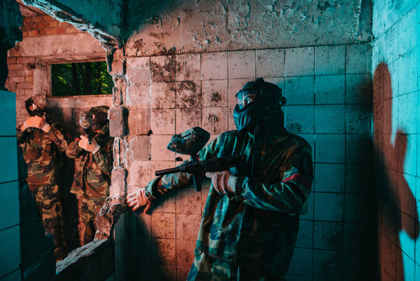 male paintball player in goggle mask and camouflage uniform hiding behind wall while other team is standing near in abandoned building  - Photo, Image
