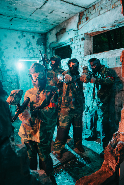 equipo de paintball en uniforme y máscaras protectoras apuntando con pistolas de paintball a cámara en edificio abandonado
 - Foto, imagen