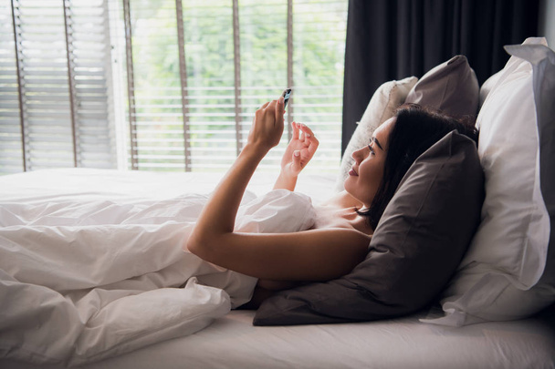 Joven mujer conectar la red social con el teléfono inteligente por la mañana, se relaja en la cama blanca
 - Foto, imagen