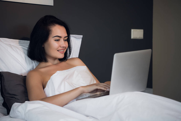 Νεαρή γυναίκα στο σπίτι κάθεται στο κρεβάτι ξύπνησε περιήγηση φορητό υπολογιστή - Φωτογραφία, εικόνα