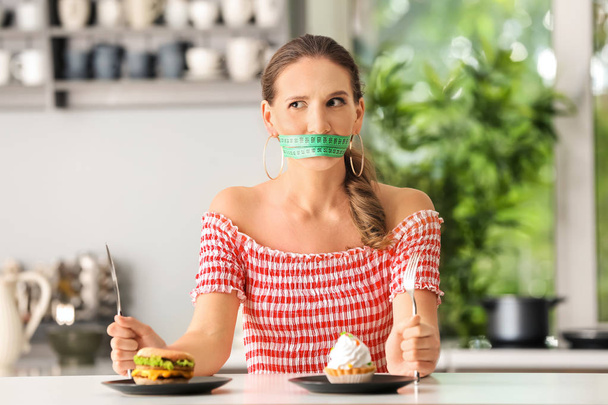 Emotional woman with measuring tape around her mouth and unhealthy food in kitchen. Diet concept - Фото, зображення