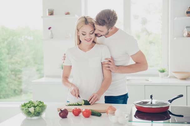 Two nice adorable positive stylish trendy sweet married people, spouses, bearded husband and blonde wife cooking dish, cutting cucumber, guy hugging girl, date, white light interior - Photo, image