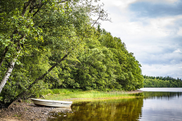 Βάρκα στην όχθη ενός ποταμού περιβάλλεται από ένα δάσος με πράσινα δέντρα, σε μια ήρεμη λίμνη - Φωτογραφία, εικόνα
