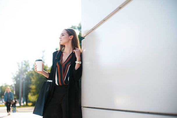 Young attractive woman exits from shopping mall with glass of coffee and package. Sale and business concept - Photo, image