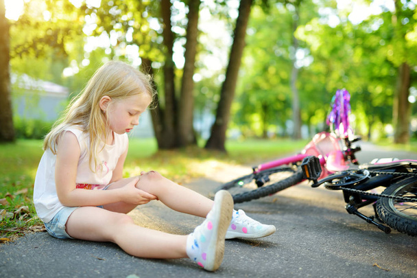 Linda niña sentada en el suelo después de caerse de su bicicleta en el parque de verano. Un niño que se lastima mientras monta en bicicleta. Activo ocio familiar con niños
. - Foto, Imagen