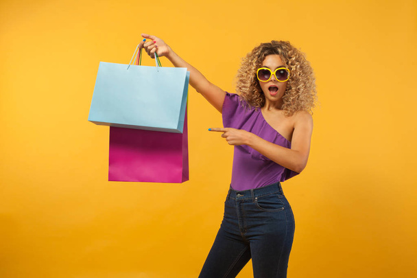 Smiling beautiful young woman with bags. Girl with afro hairstyle and sunglasses. Yellow background. Concept for sale. - Photo, Image