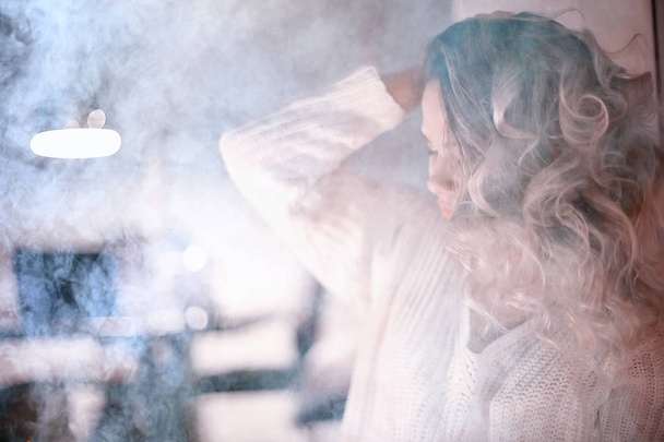 young blonde woman in white knitted sweater posing in dark room with smoke - Fotografie, Obrázek