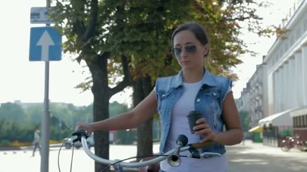 Portrait of a young, happy girl who walks around the city with a bicycle and drinks coffee - Video, Çekim