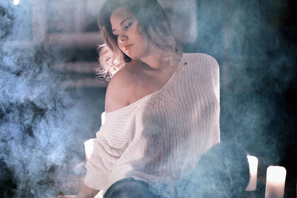 young blonde woman in white knitted sweater posing in dark room with smoke - Photo, Image