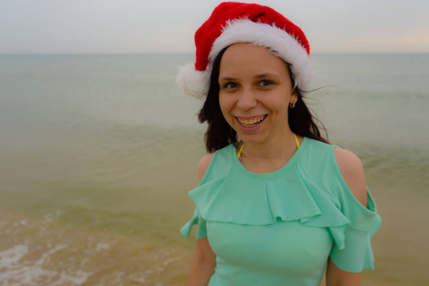 Happy girl smiling in santa claus hat on sea beach. Woman with long brunette hair posing on blurred natural background. Vacation and holidays. Seascape and nature. Christmas and new year concept. - Foto, afbeelding