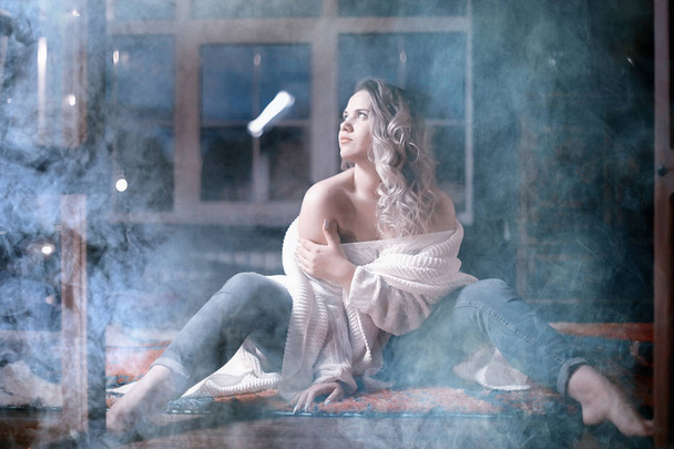 young blonde woman in white knitted sweater posing in dark room with smoke - Фото, изображение