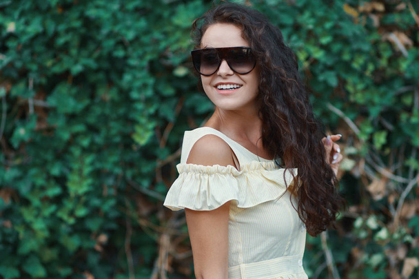 Retrato de cerca de una hermosa mujer caucásica con una sonrisa encantadora caminando, posando al aire libre, sobre el fondo de las plantas. Modelo con gafas de sol con estilo
.  - Foto, Imagen