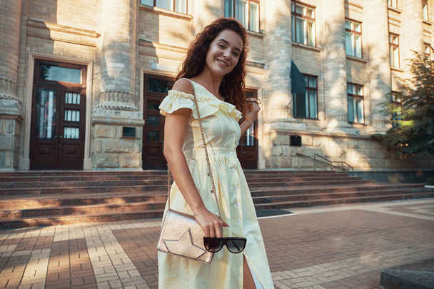 Portrait of inspired lady spending vacation in Europe. Outdoor photo of smiling, cheerful girl with brown curly hair, sunglasses, light yellow summer dress, walking around city in the morning. - Photo, Image