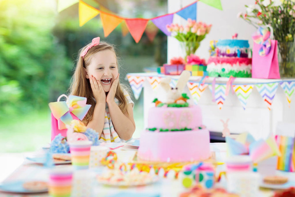 Kids birthday party with colorful rainbow pastel decoration and bunny layer cake. Little girl with sweets, candy and fruit. Balloons and banner at festive decorated table for child or baby birthday. - Photo, Image