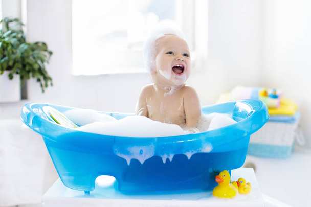 Happy laughing baby taking a bath playing with foam bubbles. Little child in a bathtub. Smiling kid in bathroom with colorful toy duck. Infant washing and bathing. Hygiene and care for young children. - Fotó, kép