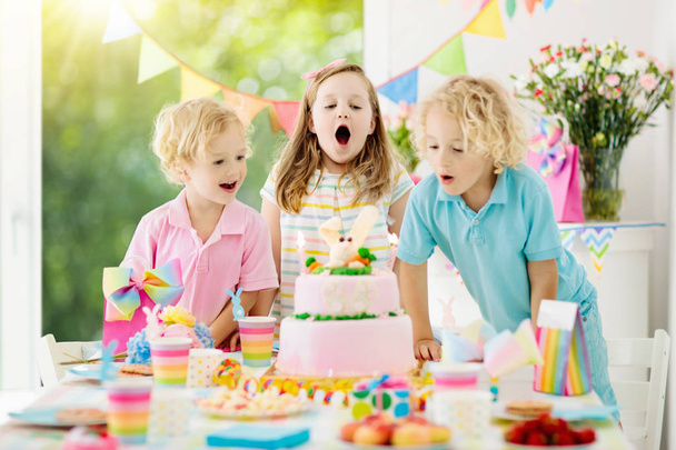 День рождения детей. Дети задувают свечи на розовом кроличьем торте. Пастельное радужное украшение и сервировка стола для детского мероприятия, баннера и флага. Девочка и мальчик с подарками на день рождения. Семейный праздник
. - Фото, изображение