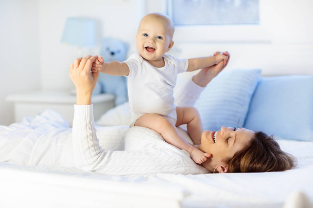 Anya és a gyermek a fehér ágy. Anya és a baba fiú pelenka lejátszása napfényes hálószoba. Szülő és pihentető otthon kis gyerek. Családi szórakozás együtt. Ágynemű és textil csecsemő gyerekszobába. - Fotó, kép