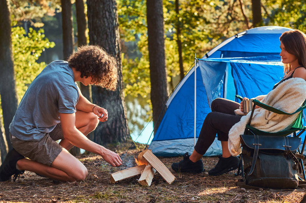Joven pareja de excursionistas en el campamento en el bosque. Joven intenta encender un fuego
.  - Foto, imagen