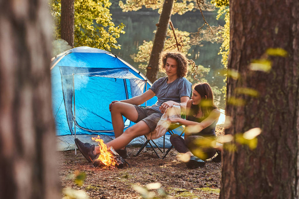 Mladý pár sedí turisté oteplování u táboráku v táboře v lese na slunečný den. - Fotografie, Obrázek