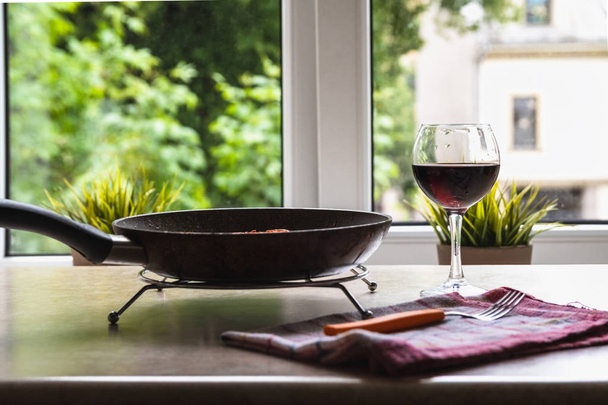 Тефлоновая сковородка и бокал вина на столе у окна
 - Фото, изображение