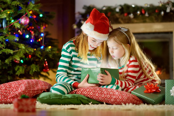 Hermanitas felices con pijamas de Navidad abriendo cajas de regalo junto a una chimenea en una acogedora sala de estar oscura en la víspera de Navidad. Celebrando la Navidad en casa
. - Foto, imagen