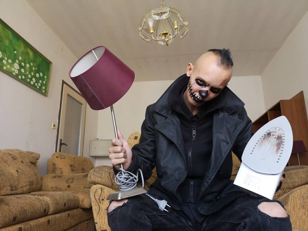 Человек со страшным макияжем на Хэллоуин с настольной лампой и утюгом в руках, сидящим в кресле  - Фото, изображение