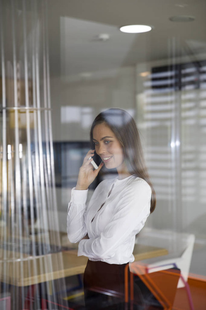 Красивая молодая женщина использует мобильный телефон за стеклом в офисе
 - Фото, изображение