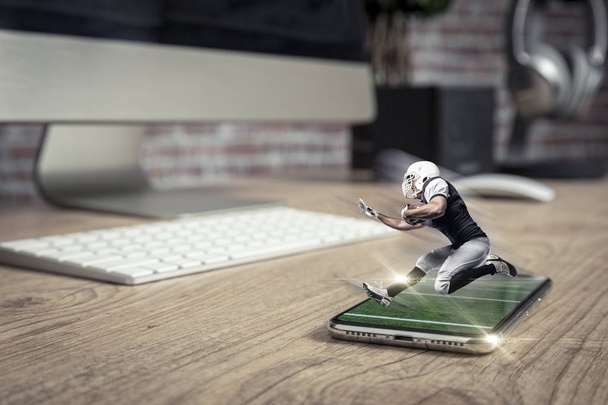 Football Player with a black uniform playing and coming out of a full screen phone on a wooden table. Watching a football game on demand concept. copy space. - Fotoğraf, Görsel