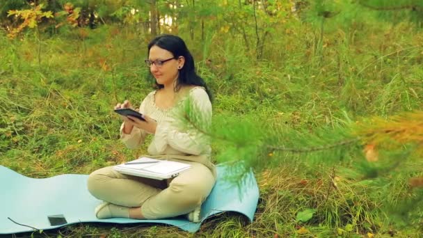 A woman in glasses at the edge of the forest works with a tablet. - Video
