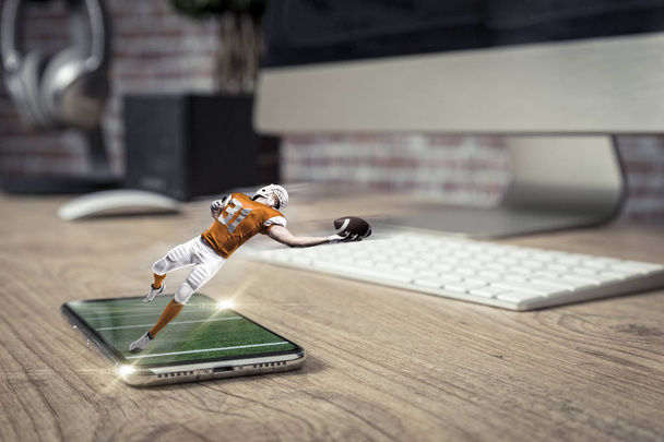 A focista egy narancssárga egységes játék, és jön ki a teljes képernyő telefon, egy fából készült asztal. Néz egy focimeccsen a követelés fogalmát. másol hely. - Fotó, kép