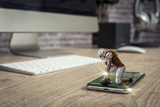Football Player with a orange uniform playing and coming out of a full screen phone on a wooden table. Watching a football game on demand concept. copy space. - Fotó, kép