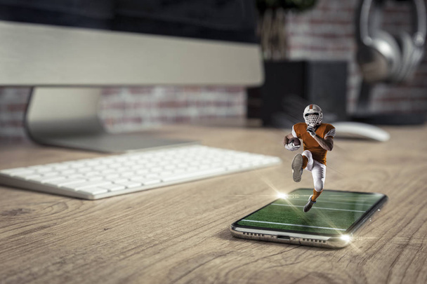 Football Player with a orange uniform playing and coming out of a full screen phone on a wooden table. Watching a football game on demand concept. copy space. - Fotoğraf, Görsel