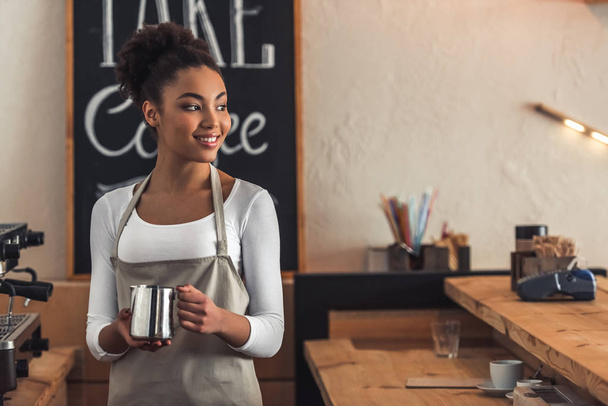 Красивая афро-американская бариста в фартуке держит чашку молока, смотрит в сторону и улыбается, стоя рядом с кофеваркой
 - Фото, изображение