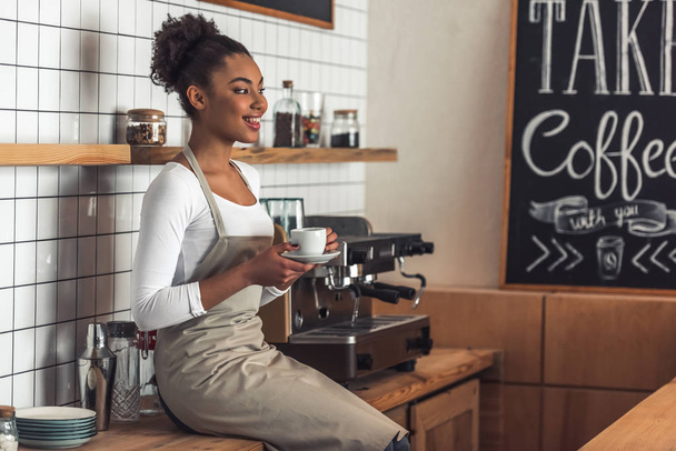 Beautiful Afro American barista in apron is holding a cup of coffee, looking away and smiling while sitting on the counter near a coffee machine - Photo, Image