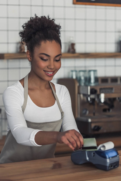 Beautiful Afro American barista in apron is using a payment terminal and smiling while standing at bar counter - Photo, Image