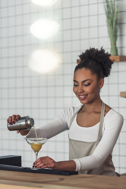 Красивая афро-американская бариста в фартуке готовит коктейль в шейкере и улыбается стоя у барной стойки
 - Фото, изображение