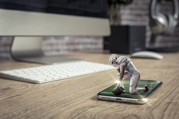 Football Player with a white uniform playing and coming out of a full screen phone on a wooden table. Watching a football game on demand concept. copy space. - Zdjęcie, obraz