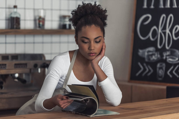 Szép Afro-amerikai unatkozni barista kötény olvasó egy magazin támaszkodva a bárban, míg a számláló - Fotó, kép