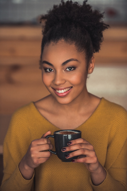 Beautiful Afro American girl in casual clothes is holding a cup of coffee, looking at camera and smiling while resting in cafe - Photo, Image