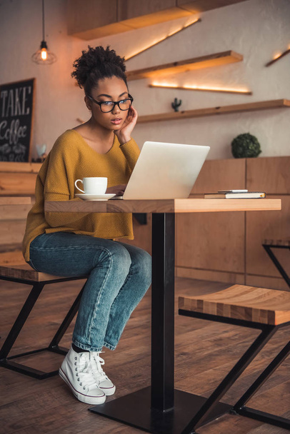 Beautiful Afro American girl in casual clothes and glasses is working with a laptop in cafe - Photo, Image
