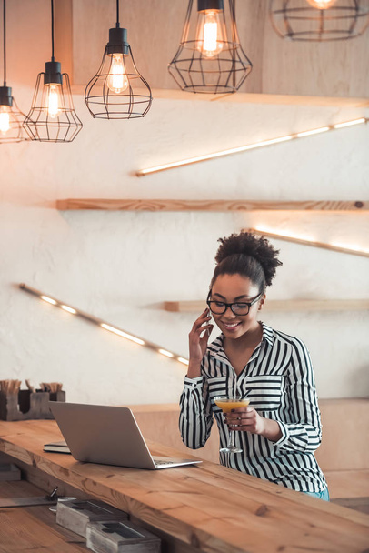 Menina afro-americana bonita em roupas casuais inteligentes e óculos está falando no telefone celular, bebendo coquetel e sorrindo enquanto trabalhava com um laptop no café
 - Foto, Imagem