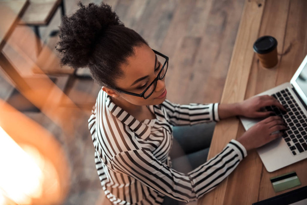 Vue grand angle de la belle afro-américaine en vêtements décontractés intelligents et lunettes de travail avec un ordinateur portable dans le café
 - Photo, image